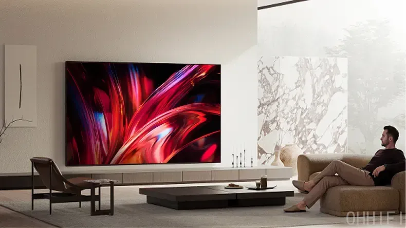 Sony Bravia 9 II on a living room wall showing a red abstract swirl, as a man watches from a beige sofa nearby