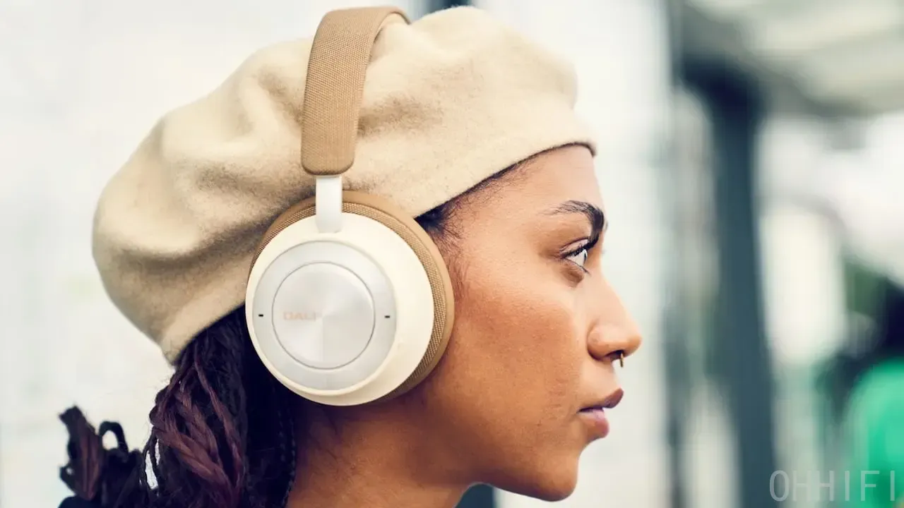 Woman wearing DALI IO-8 wireless headphones in Caramel White outdoors, paired with a beige beret, shown from the side in city surroundings.