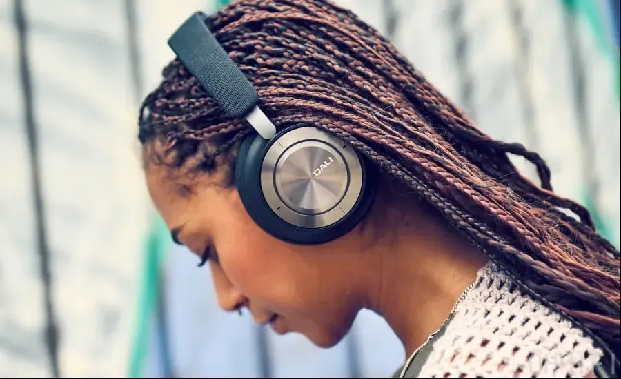 Woman wearing DALI IO-8 wireless headphones in Iron Black outdoors, head tilted down, the brushed metal ear cup with DALI logo in focus.