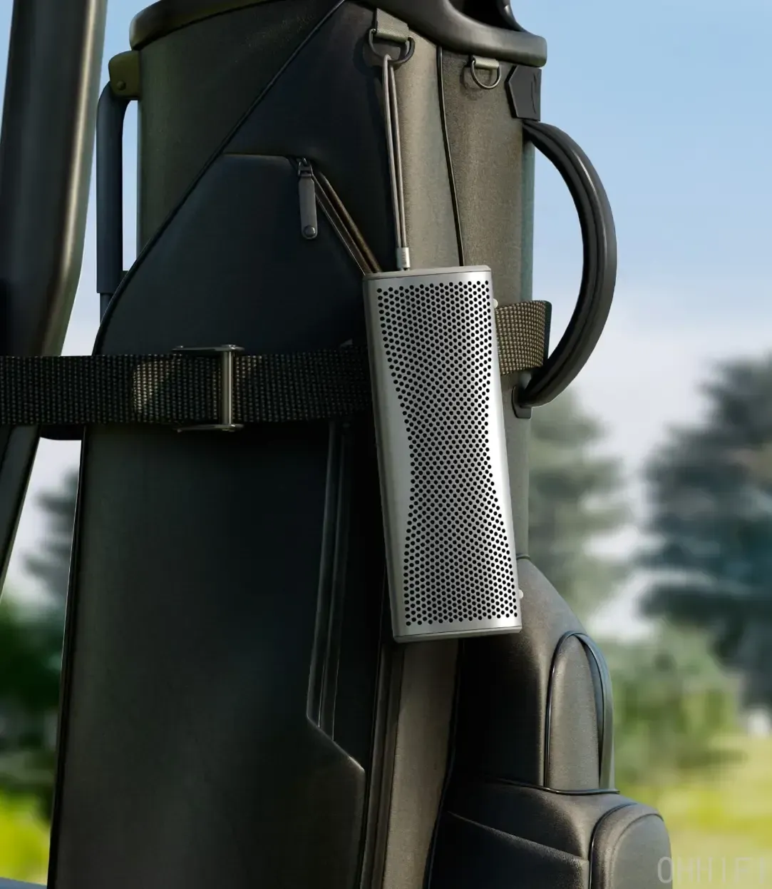 KEF Muo portable Bluetooth speaker in Silver Dusk hung by its braided strap from a black golf bag on a course, showing its perforated aluminum grille and lifestyle outdoor portability.