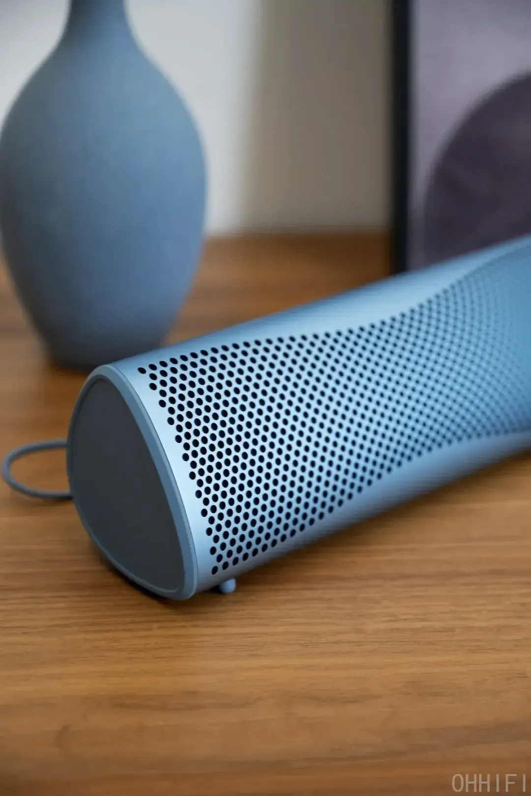 KEF Muo portable Bluetooth speaker in Blue Aura, lying flat on a wooden tabletop with its perforated aluminum grille, end cap and braided strap in focus, ceramic vase behind.