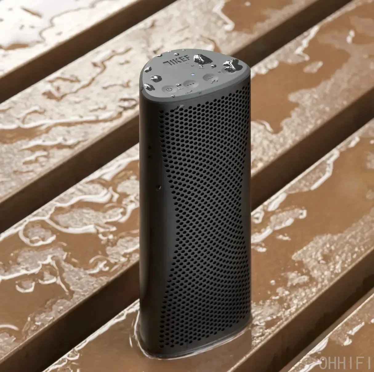 KEF Muo portable Bluetooth speaker in Midnight Black standing upright on rain-soaked wooden decking, water droplets pooled on the top control panel, demonstrating its IP67 waterproof rating.