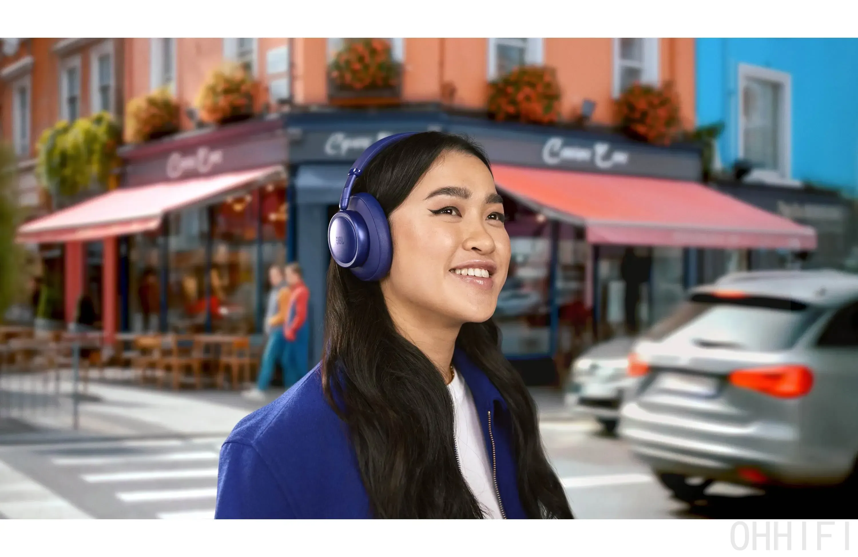 A person wearing blue JBL Live 780NC wireless headphones on a busy city street, with cafés and traffic blurred behind.