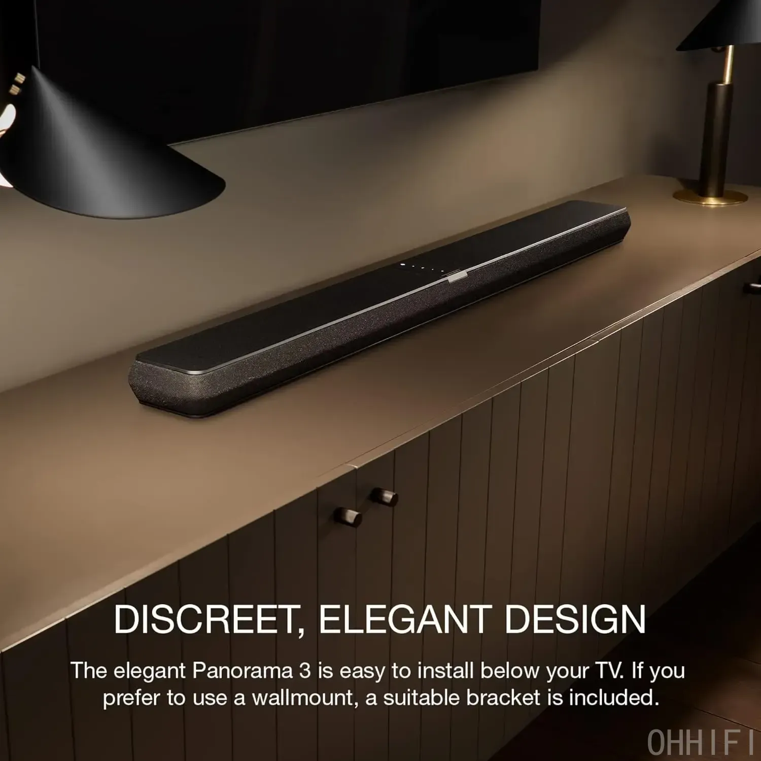 Bowers & Wilkins Panorama 3 black soundbar on a wood console beneath a wall-mounted TV in a warmly lit room with a lamp and pendant light.